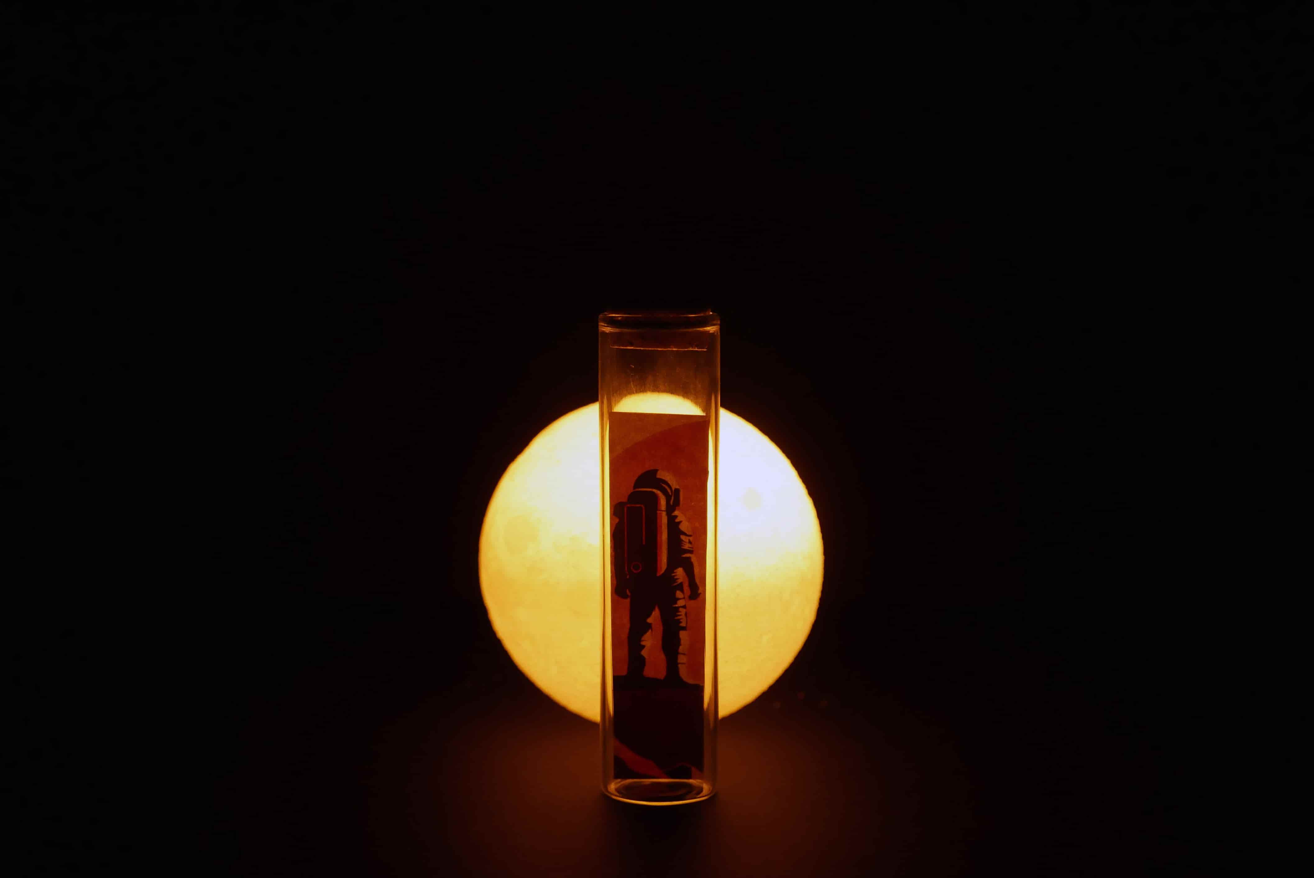 Una fotografia in still life di un disegno di un astronauta dentro una fialetta che guarda la luna rossa