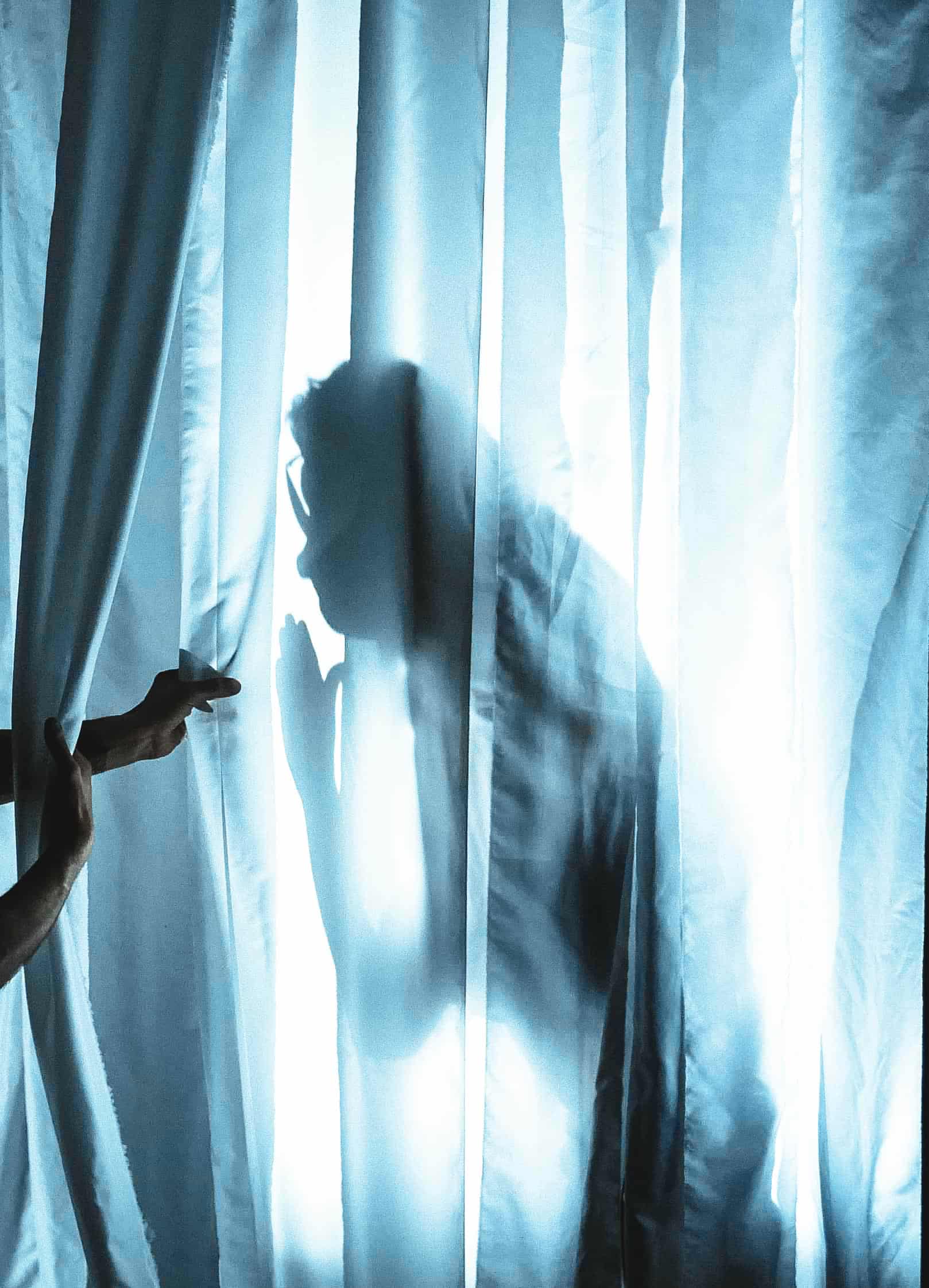 Sagoma di una persona dietro delle tende blu trasparenti, con una mano premuta contro il tessuto e l’altra che tira la tenda per aprirla sul lato sinistro, in controluce rispetto alla luce intensa.