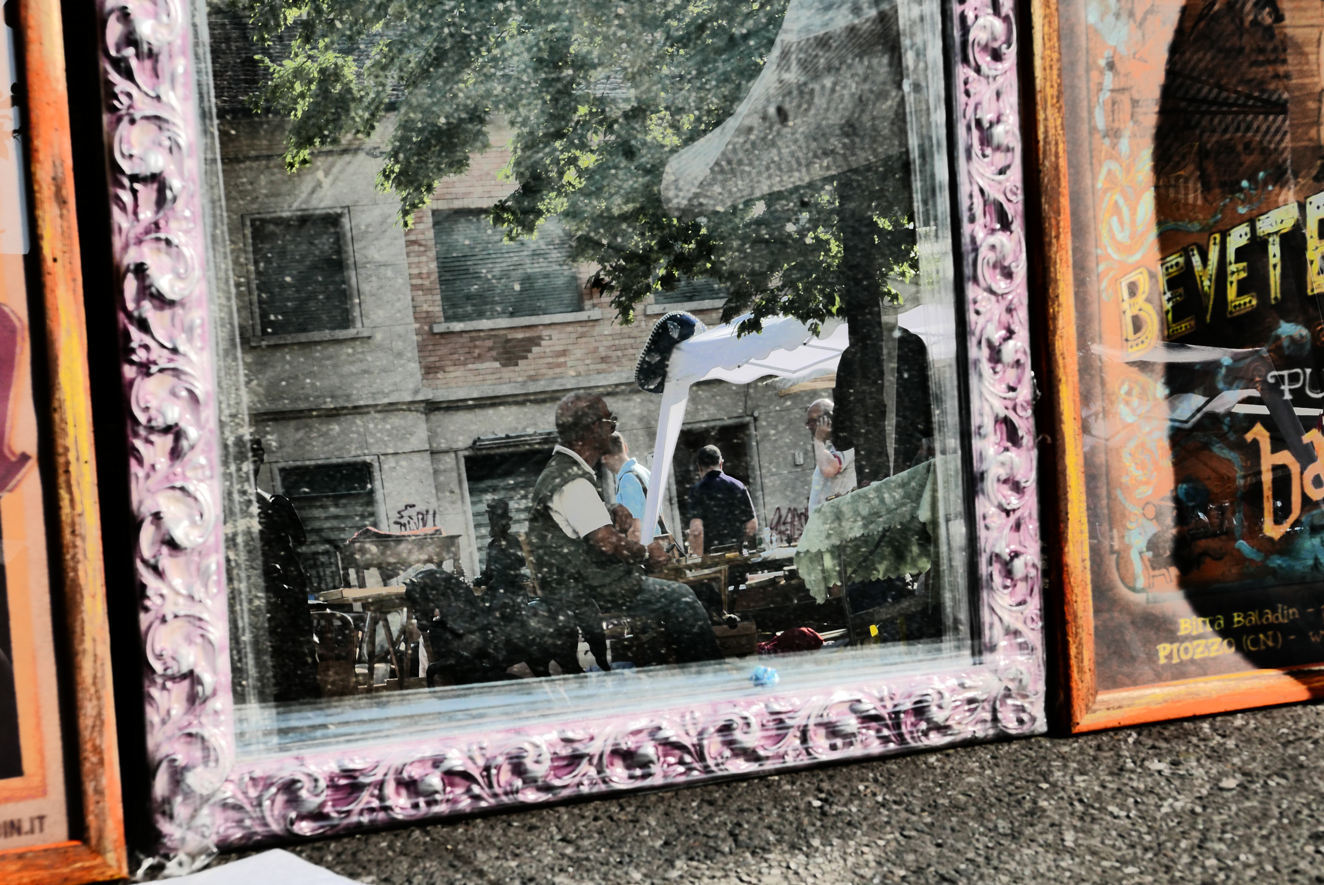 Un grande specchio decorato riflette le persone sedute ai tavoli all'aperto sotto ad alberi e tendoni,edifici fanno da sfondo sullo sfondo.