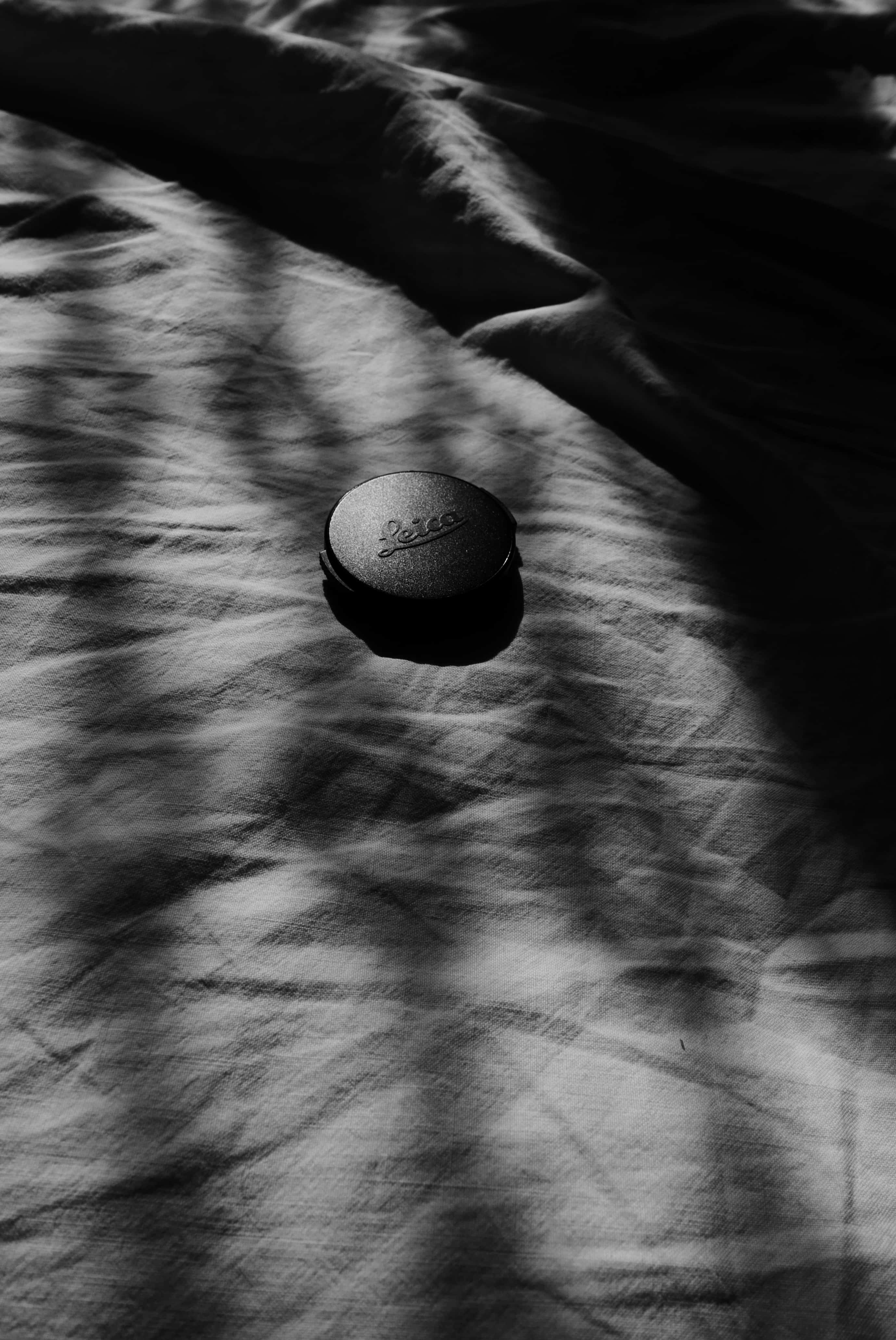 Foto in bianco e nero di un copriobiettivo Leica in primo piano su lenzuola stropicciate, con giochi di luci e ombre in diagonale.