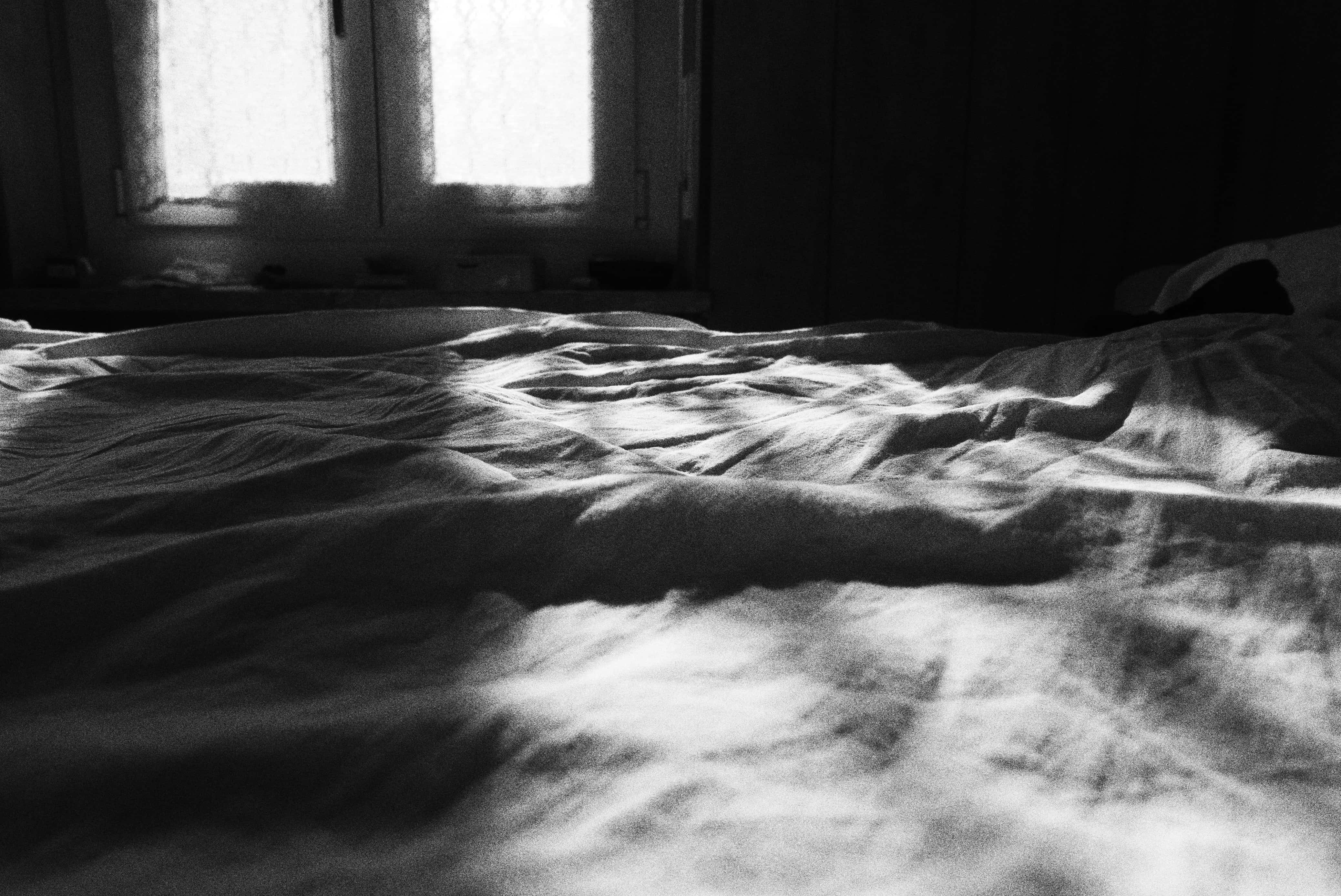 Foto in bianco e nero che ritrae un letto sfatto, con la luce del sole che filtra attraverso i vetri di due finestre sullo sfondo, creando macchie luminose e ombre sulle lenzuola.