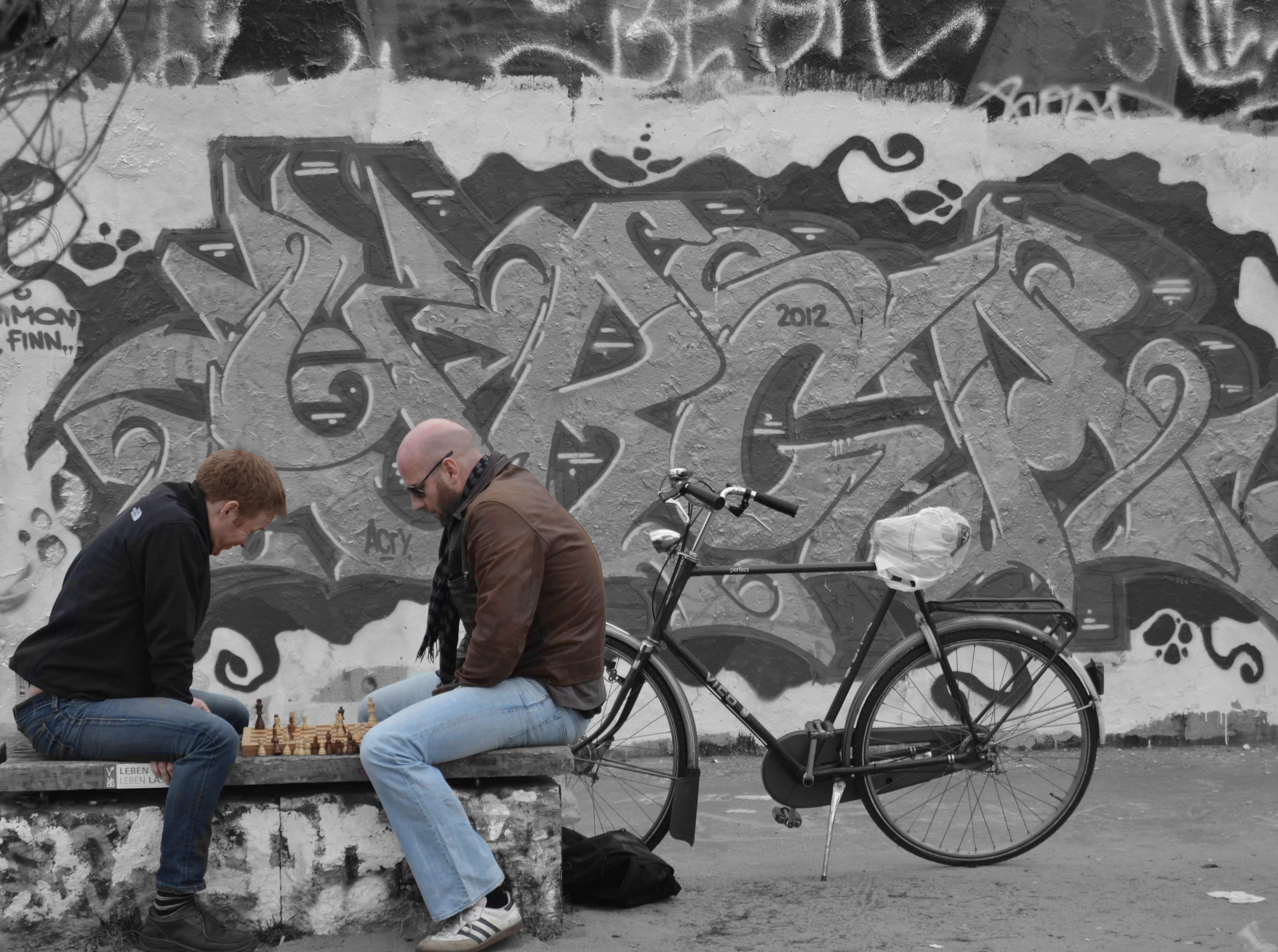 Due persone che davanti al muro di berlino giocano a scacchi.