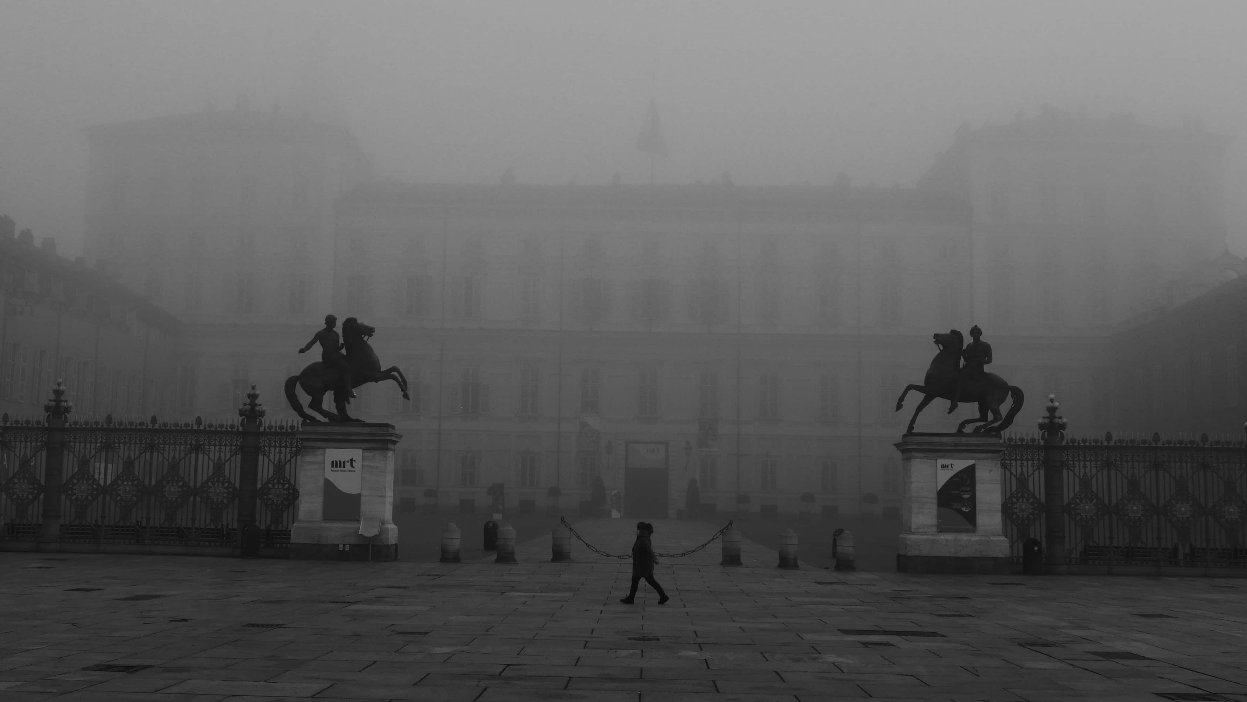 Foto in bianco e nero, in una piazza, il soggetto centrale cammina e lo sfondo è un castello avvolto nelle nebbia.